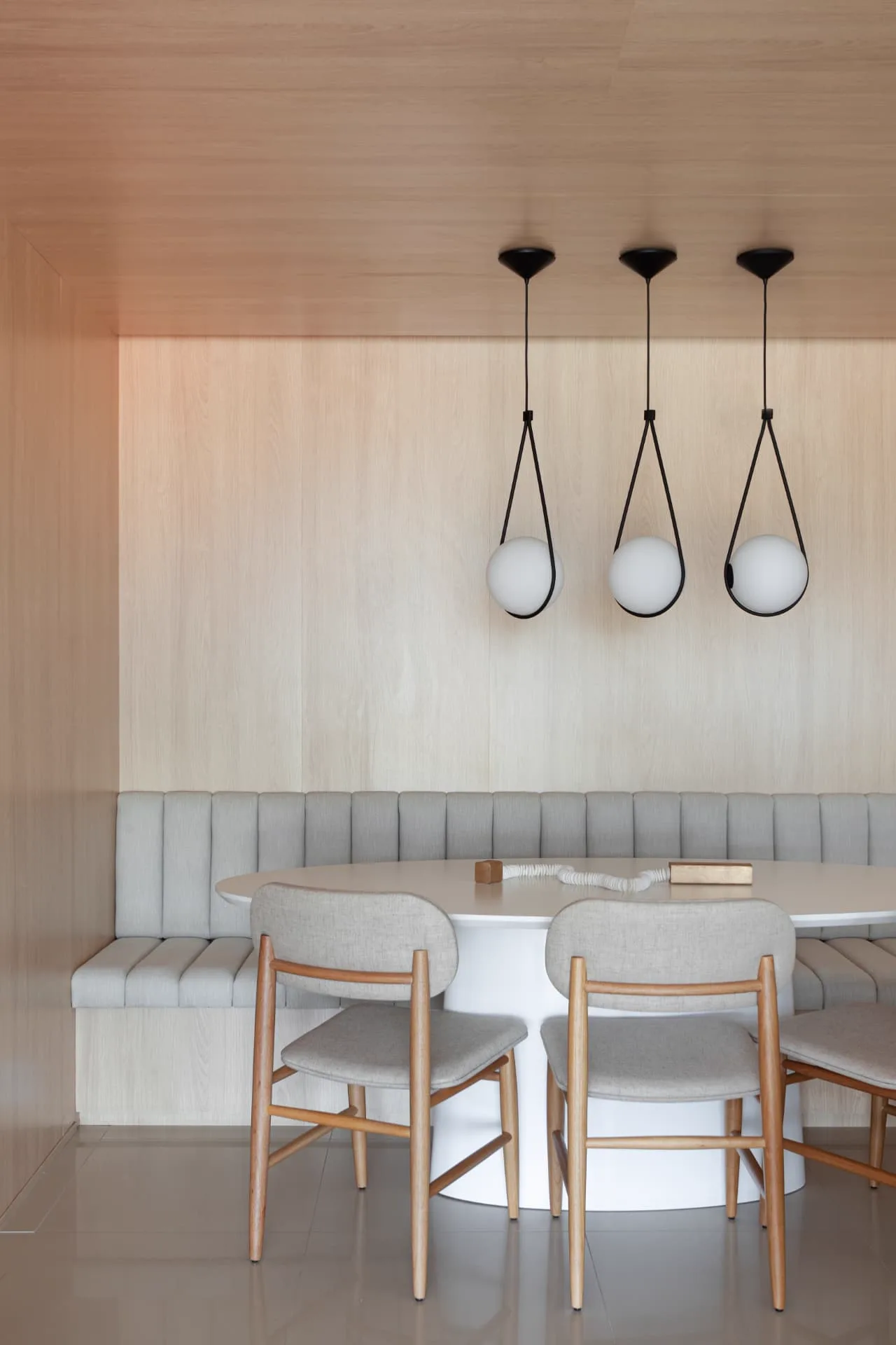 Canto de jantar com banco estofado sálvia, mesa oval branca e três pendentes esféricos