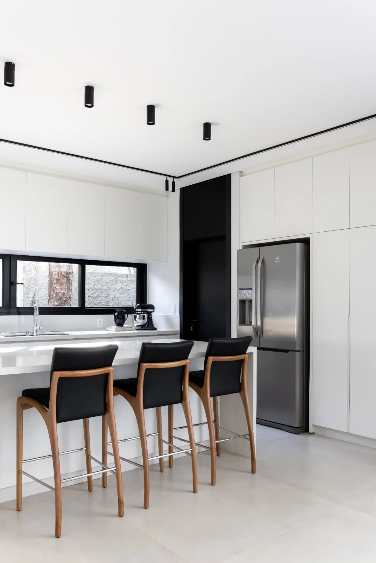 Cozinha em off white com bancada clara, banquetas de madeira e couro preto e iluminação em spots pretos