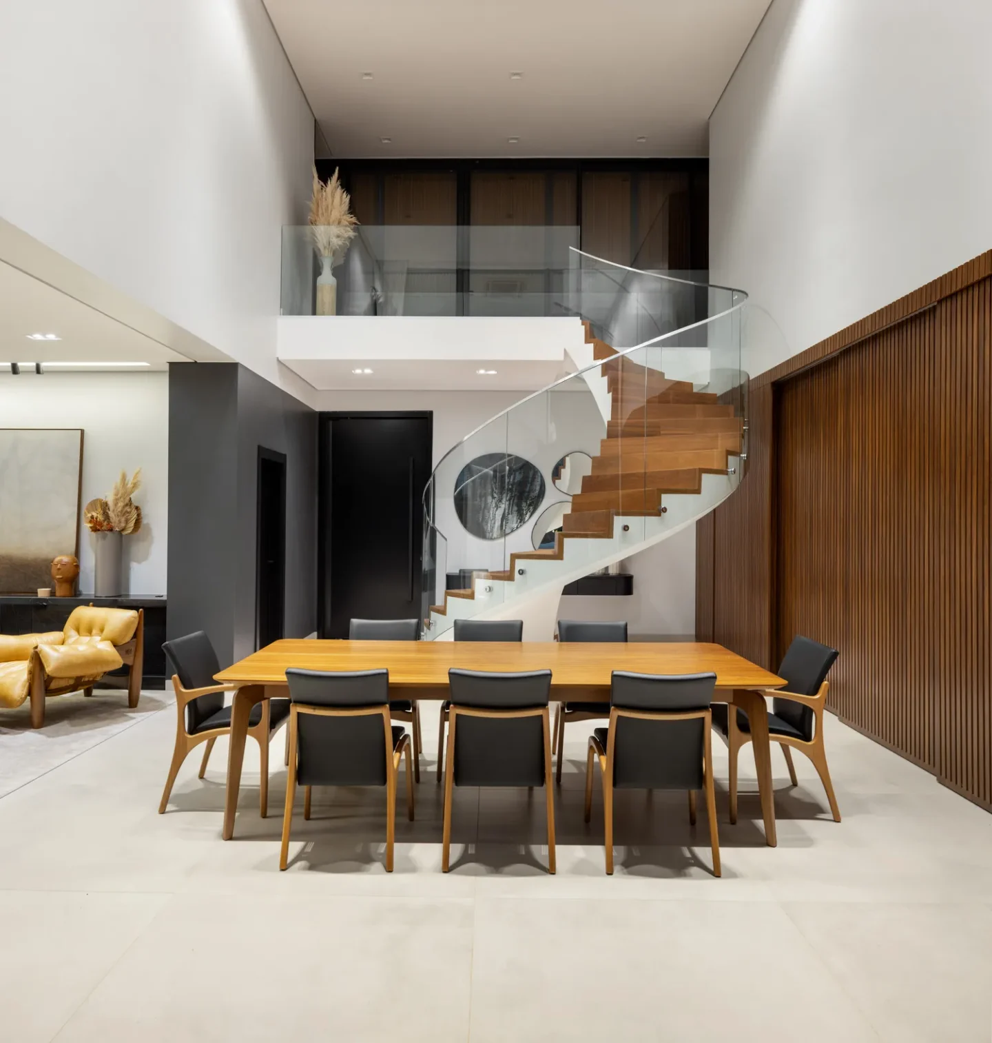 Sala de jantar com pé direito duplo, mesa de dez lugares em madeira e escada helicoidal com guarda-corpo de vidro curvo