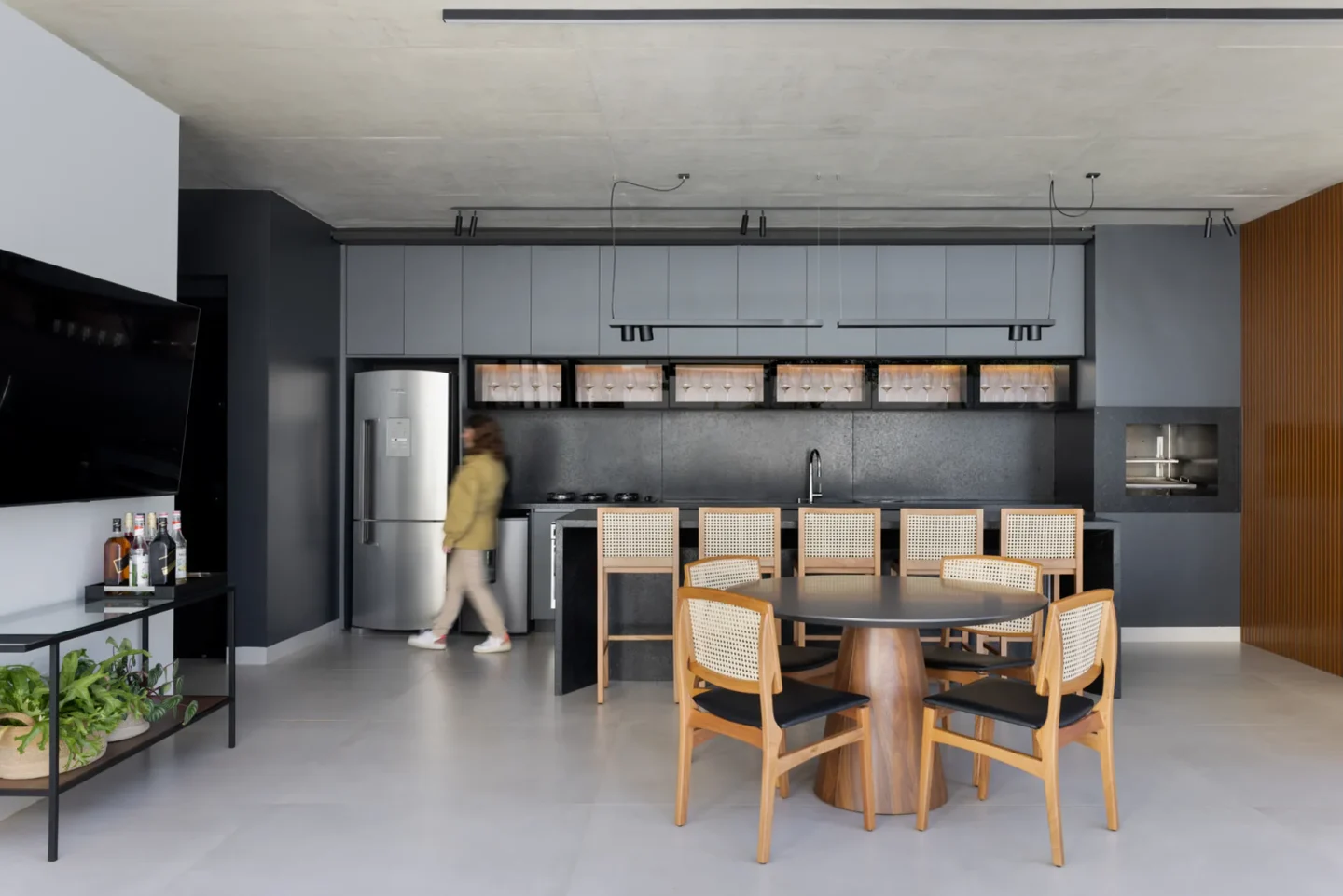 Espaço gourmet com marcenaria grafite, bancada de pedra escura, adega aberta iluminada e mesa redonda de madeira