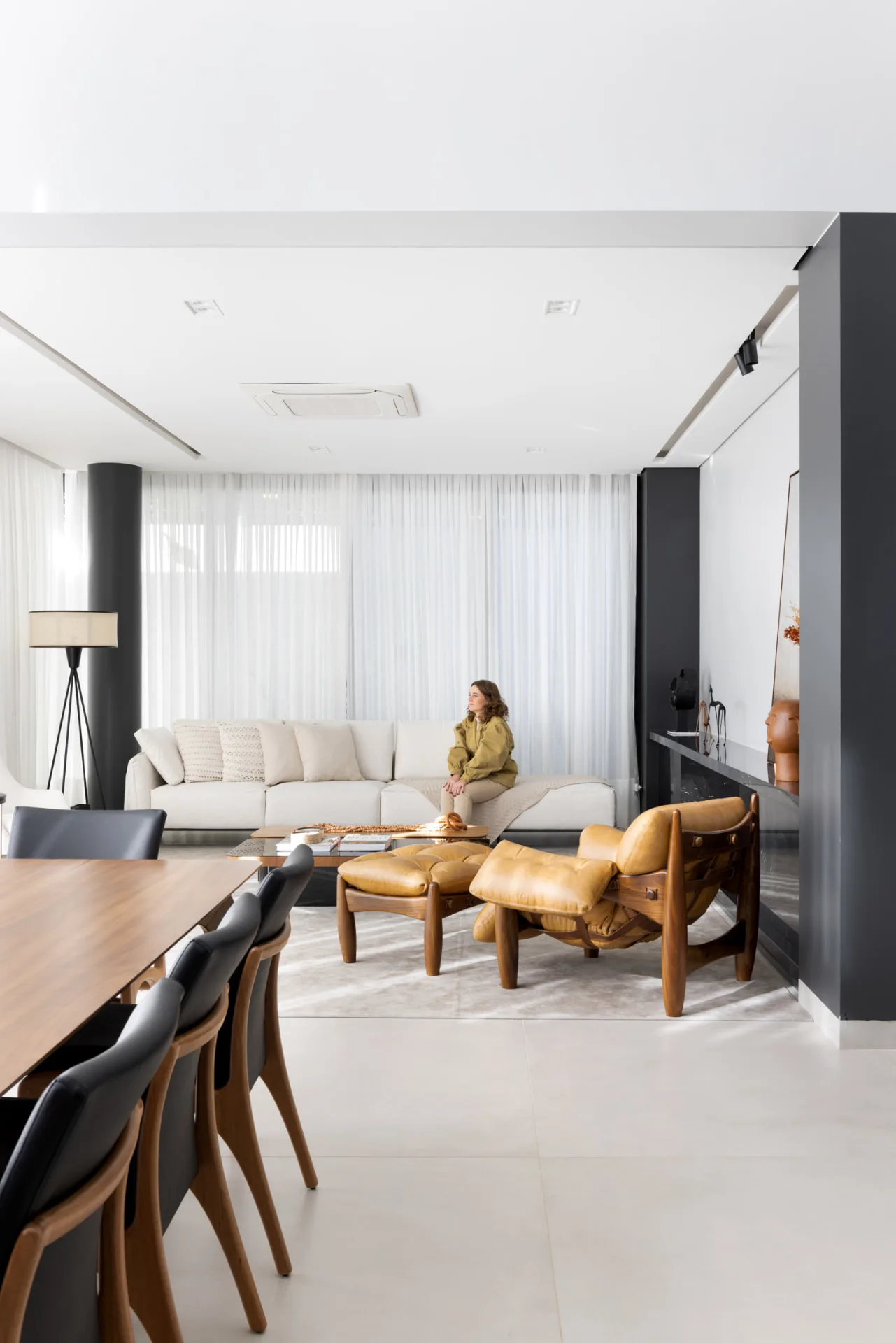 Vista da mesa de jantar para o living com cortinas de voil branco e poltrona de couro caramelo