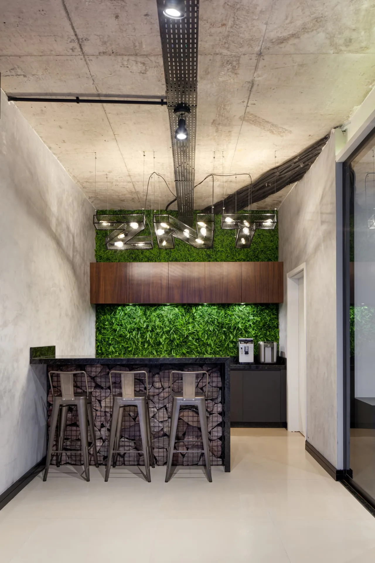 Copa com jardim preservado, nome da empresa em luminárias de metal perfurado, bancada apoiada em gabião de pedras e banquetas metálicas