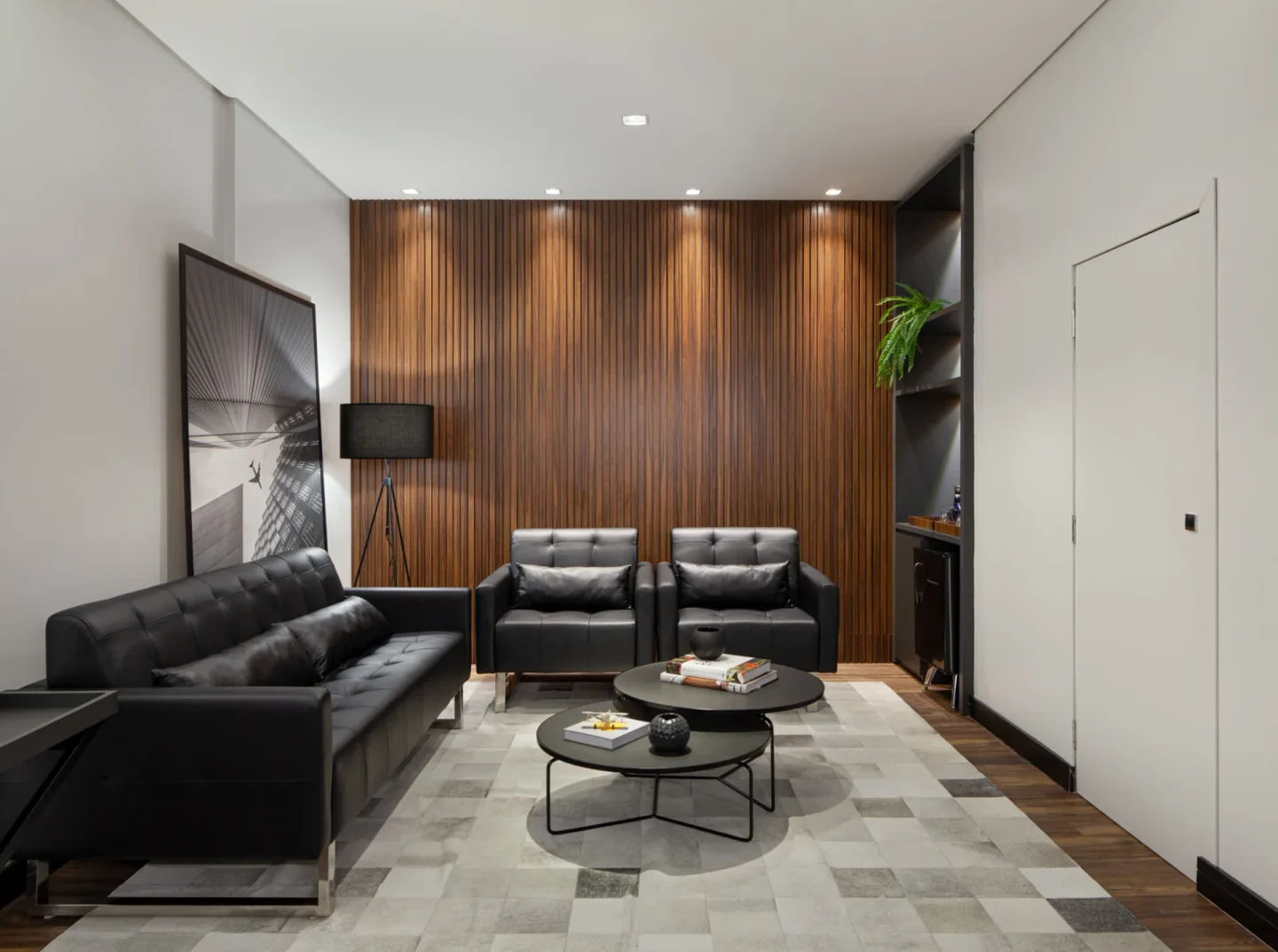 Lounge com sofás de couro preto capitonê, painel de madeira ripada, tapete de couro em tons de cinza e mesas de centro redondas