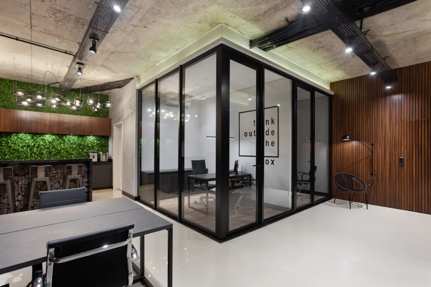 Open space do escritório com cabine de vidro e esquadria preta, laje de concreto aparente, eletrocalhas pretas e copa ao fundo