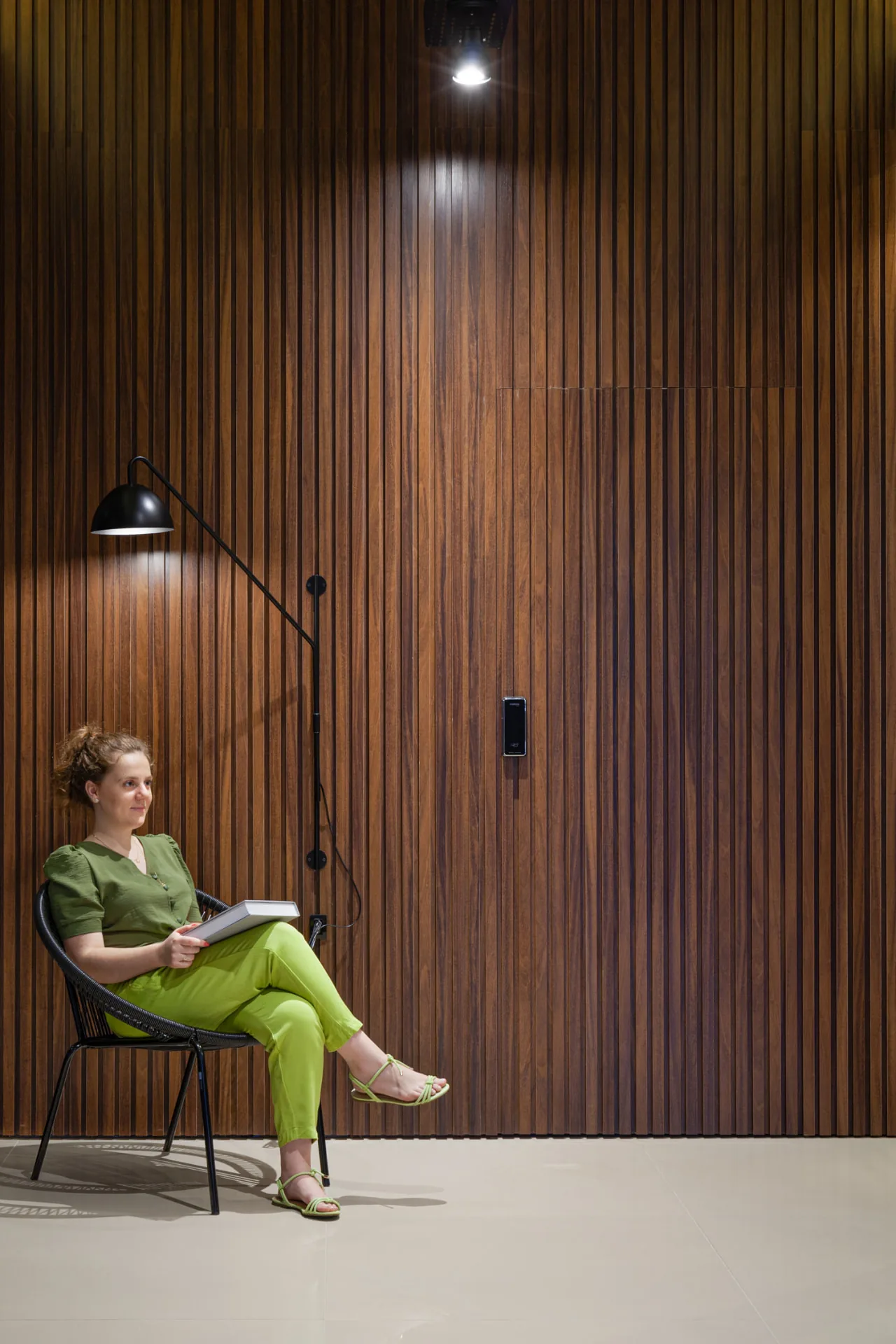 Mulher sentada em cadeira de fio preto diante do painel de madeira ripada de piso a teto, com luminária articulada de parede