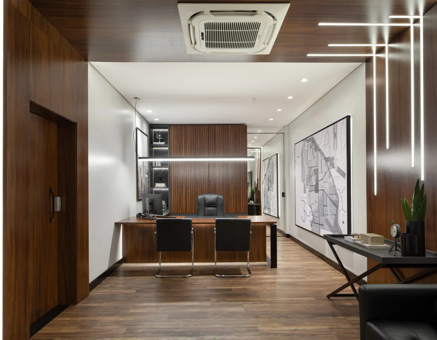 Sala da diretoria com painel ripado atrás da mesa, forro de madeira com perfis de luz e mapas da cidade emoldurados na parede