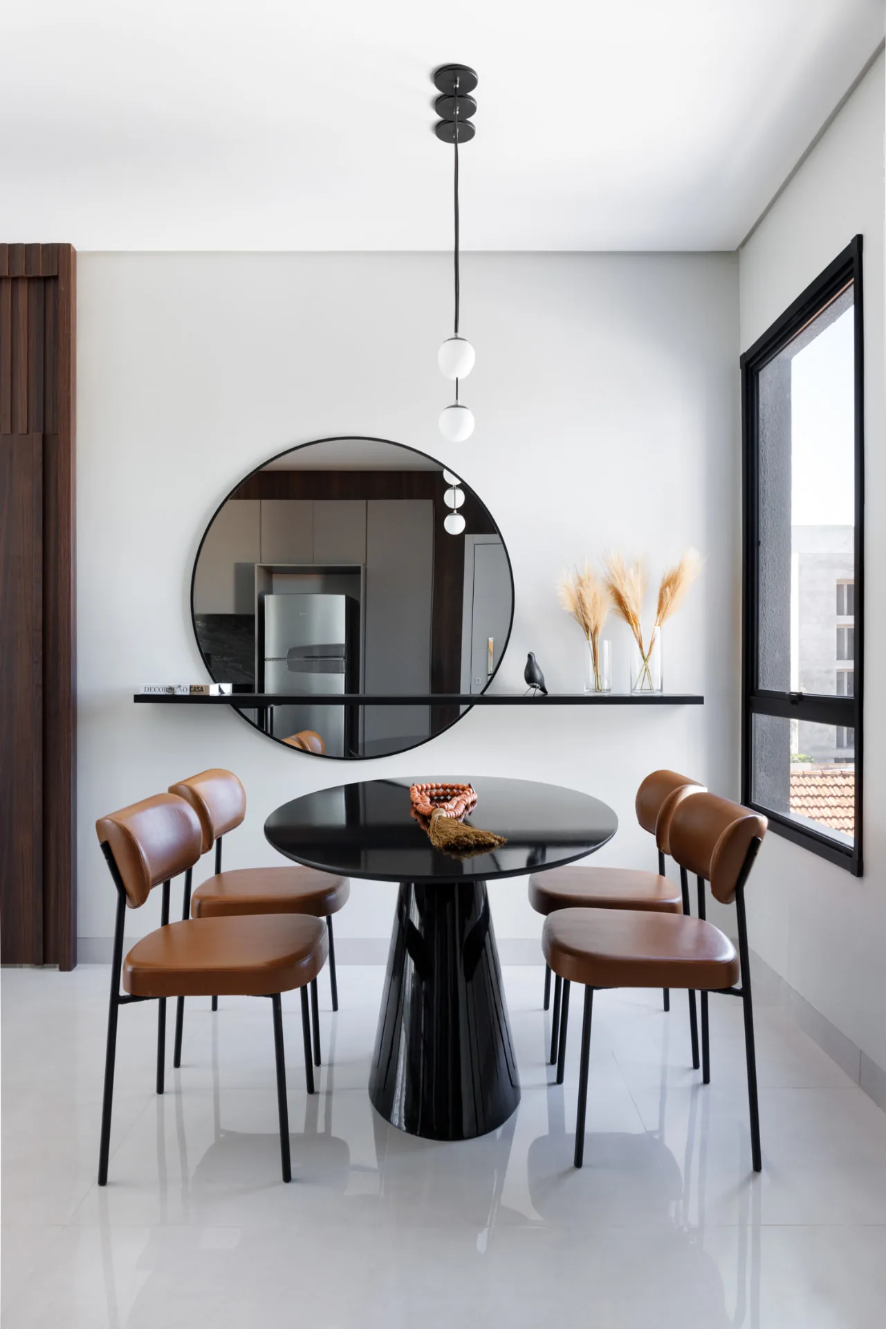 Canto de jantar com mesa redonda preta, cadeiras de couro caramelo, espelho circular e pendentes de vidro