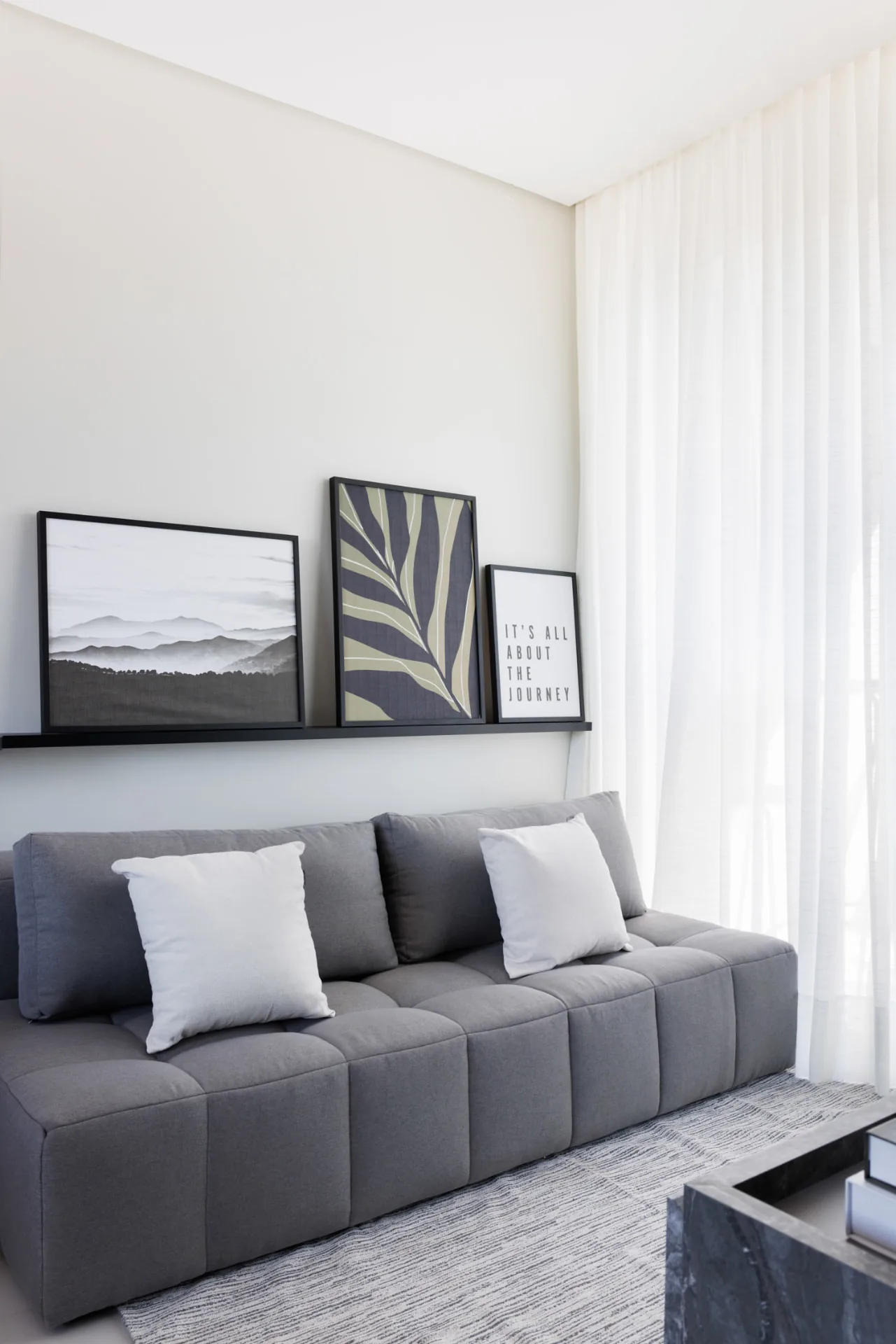 Sofá modular cinza com almofadas claras e três quadros apoiados em prateleira preta na parede branca